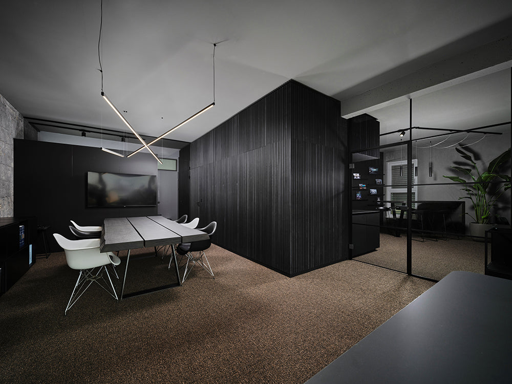 Bureau moderne équipé dun luminaire géométrique, dune longue table, de chaises blanches et noires, de murs acoustiques en bois Akupanel foncé, dune télévision et dune salle de réunion aux parois vitrées.